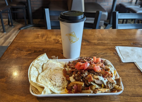 A plate of loaded nachos topped with meat, tomatoes, and two fried eggs, alongside a coffee cup on a wooden table.